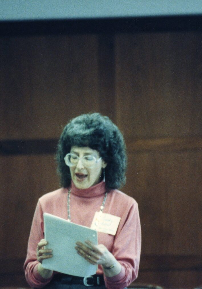 Sandy Asher reading from papers wearing a pink short sleeve shirt, poofy brown hair, and round glasses.